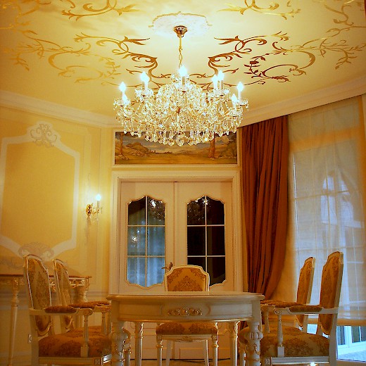 Speiseraum: Der Kronleuchter setzt das barocke Goldornament der Decke in Szene, so dass es je nach Blickwinkel matt oder glänzend schimmert